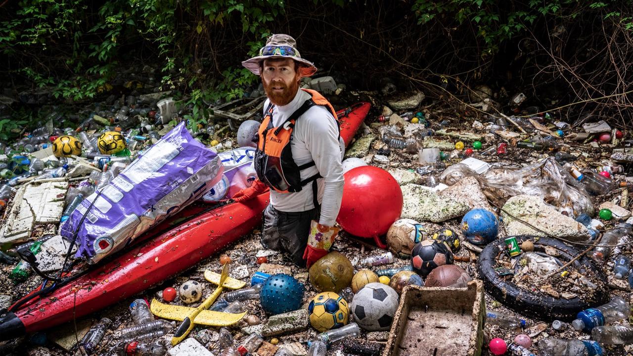 Beau Miles standing beside his red kayak in a river channel choked with plastic bottles and washed-up debris.