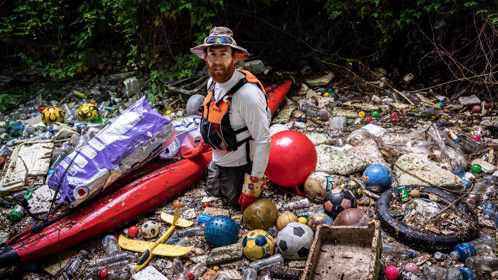 Beau Miles standing beside his red kayak in a river channel choked with plastic bottles and washed-up debris.