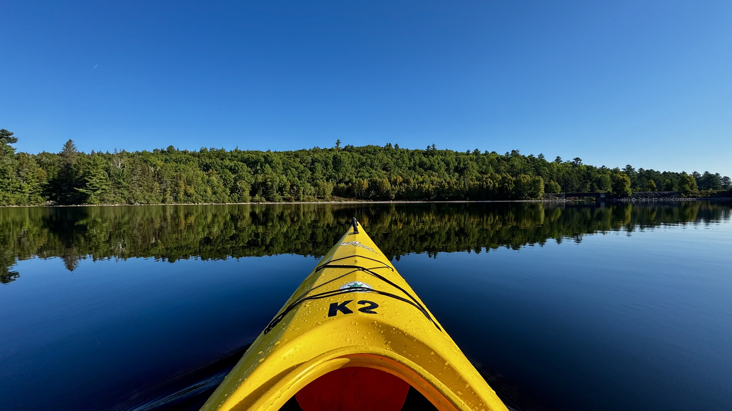 The bow of a yellow kayak points across still water toward a forested shore beneath a clear blue sky